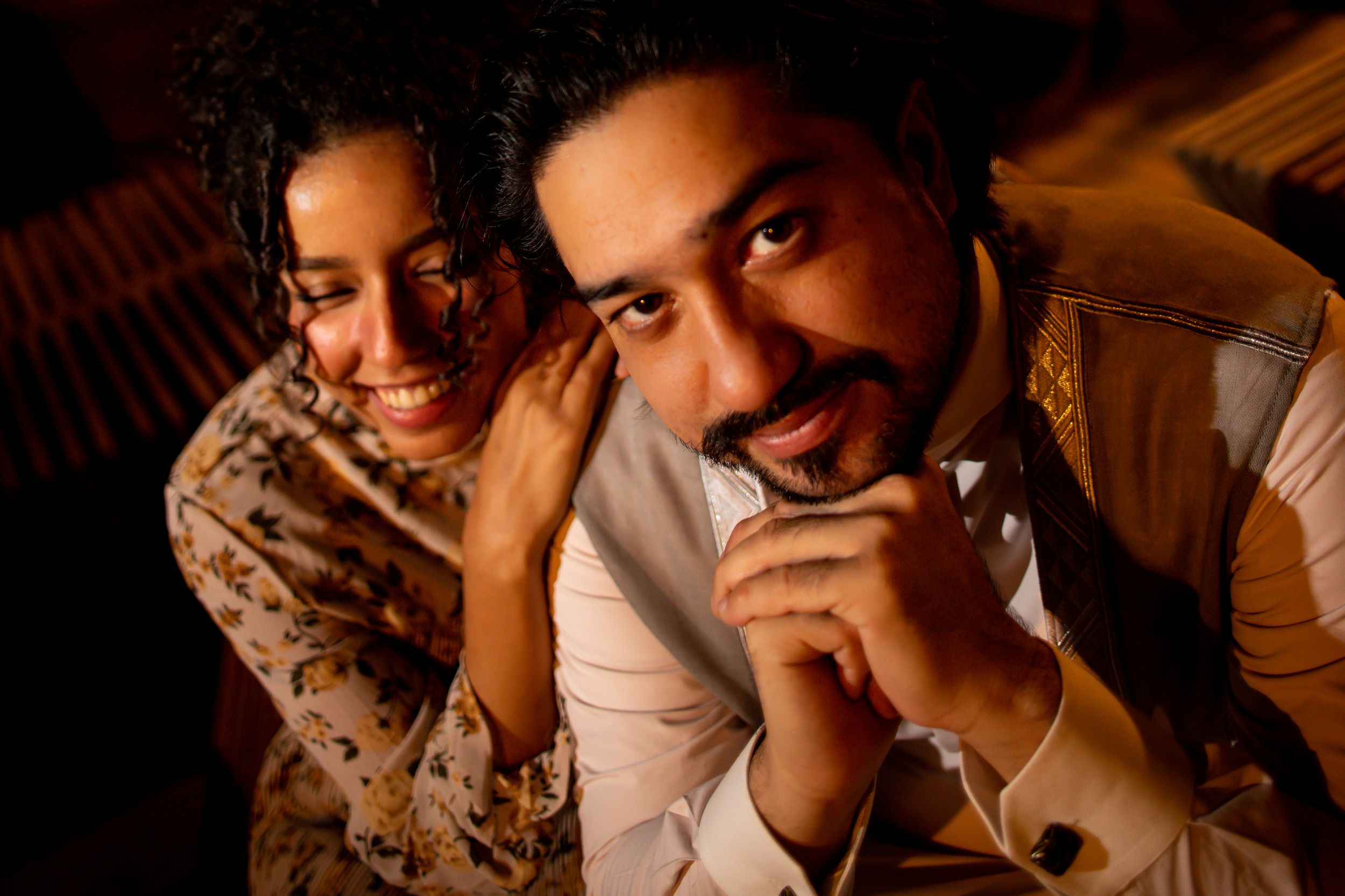 A smiling couple poses intimately in warm lighting, with a woman leaning on a man's shoulder as he clasps his hands.