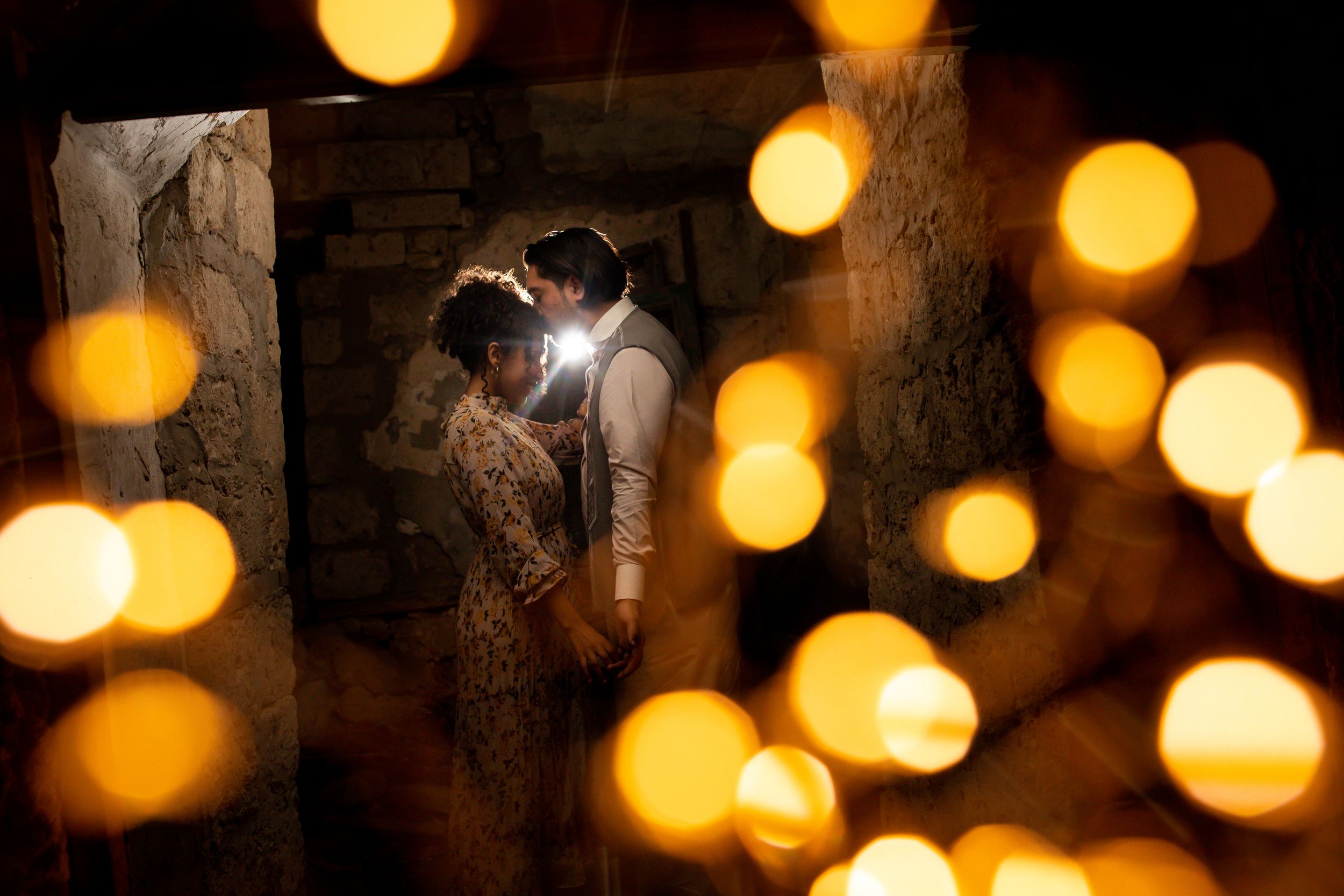 Romantic couple sharing an intimate moment surrounded by glowing bokeh candle lights in a rustic stone setting.