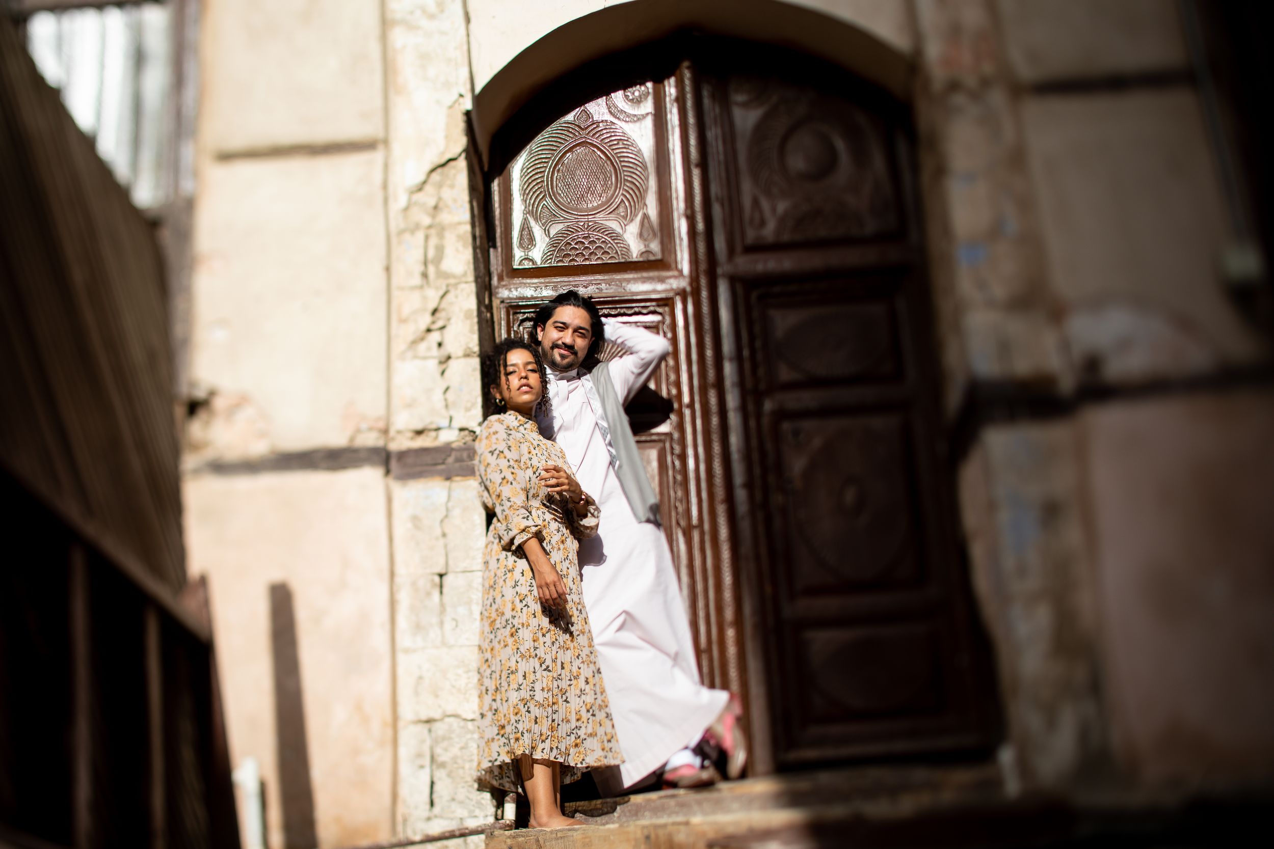 A couple poses in an ornate wooden doorway of an ancient building, dressed in traditional South Asian attire.