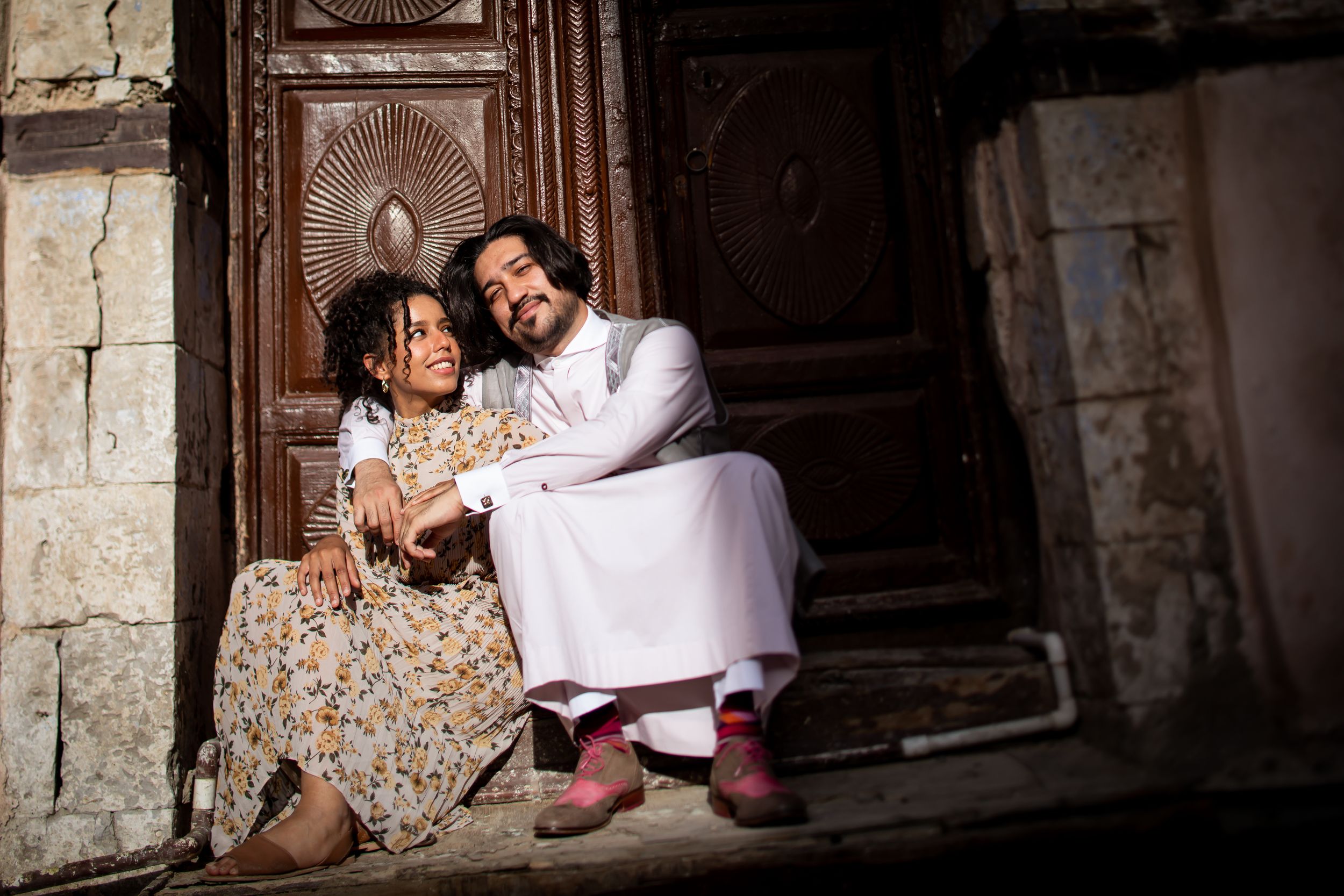 A man and young girl sit together smiling on stone steps by an ornate wooden door.
