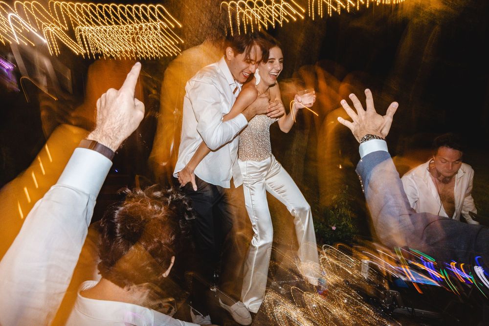 Couple dancing joyfully at wedding reception with guests, motion blur lights, bride in white jumpsuit, groom in white suit.