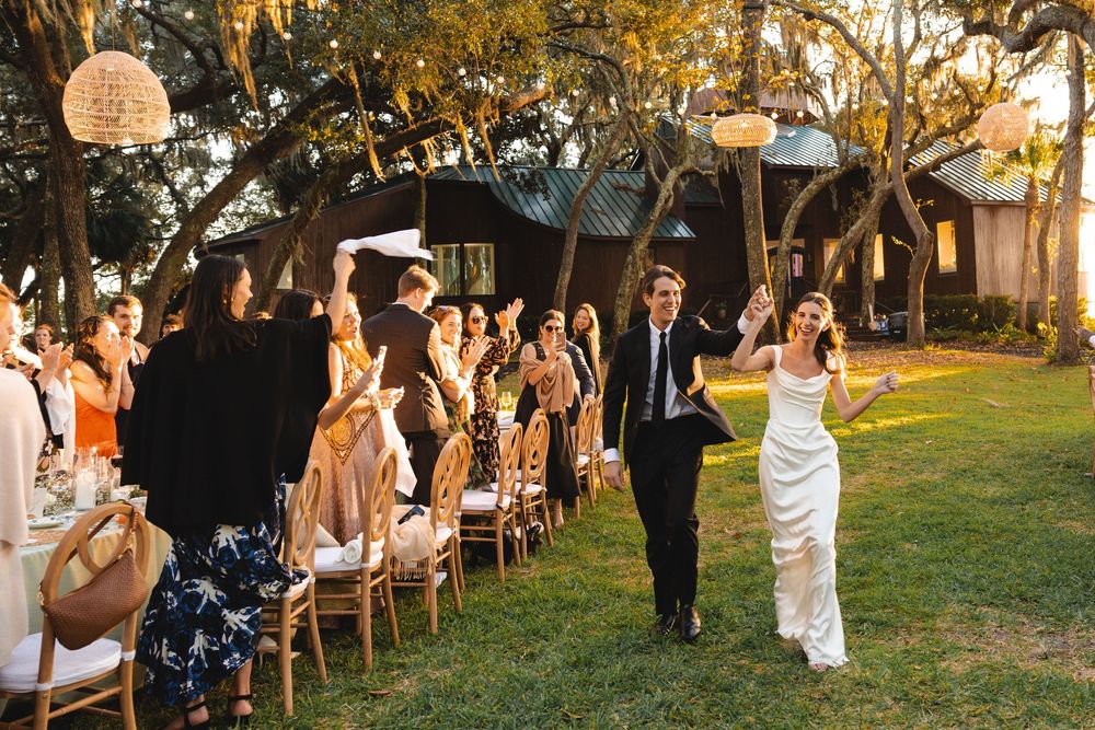 Bride and groom walk through cheering guests at an outdoor wedding reception under oak trees at golden hour.