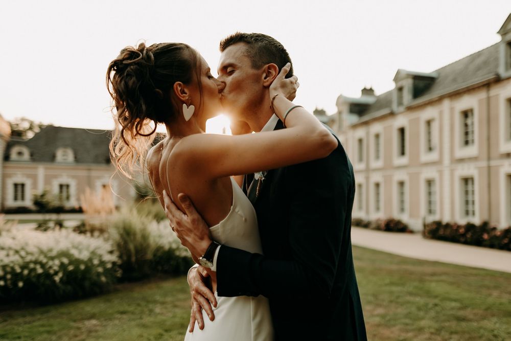 A romantic couple shares a tender kiss in a garden setting at sunset with elegant architecture in the background.