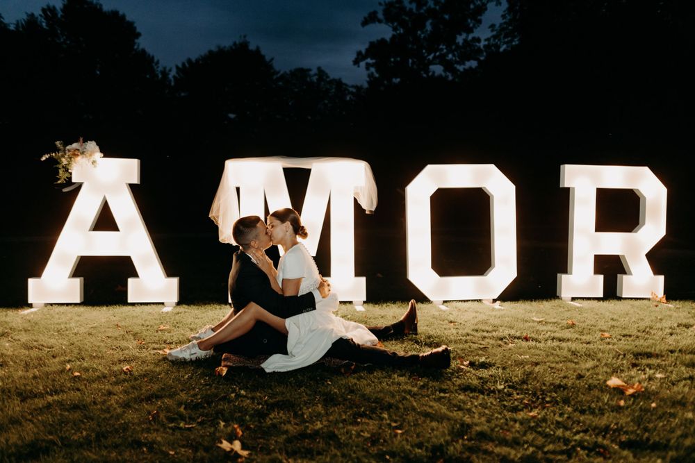 Couple seated in front of large illuminated AMOR sign at night.