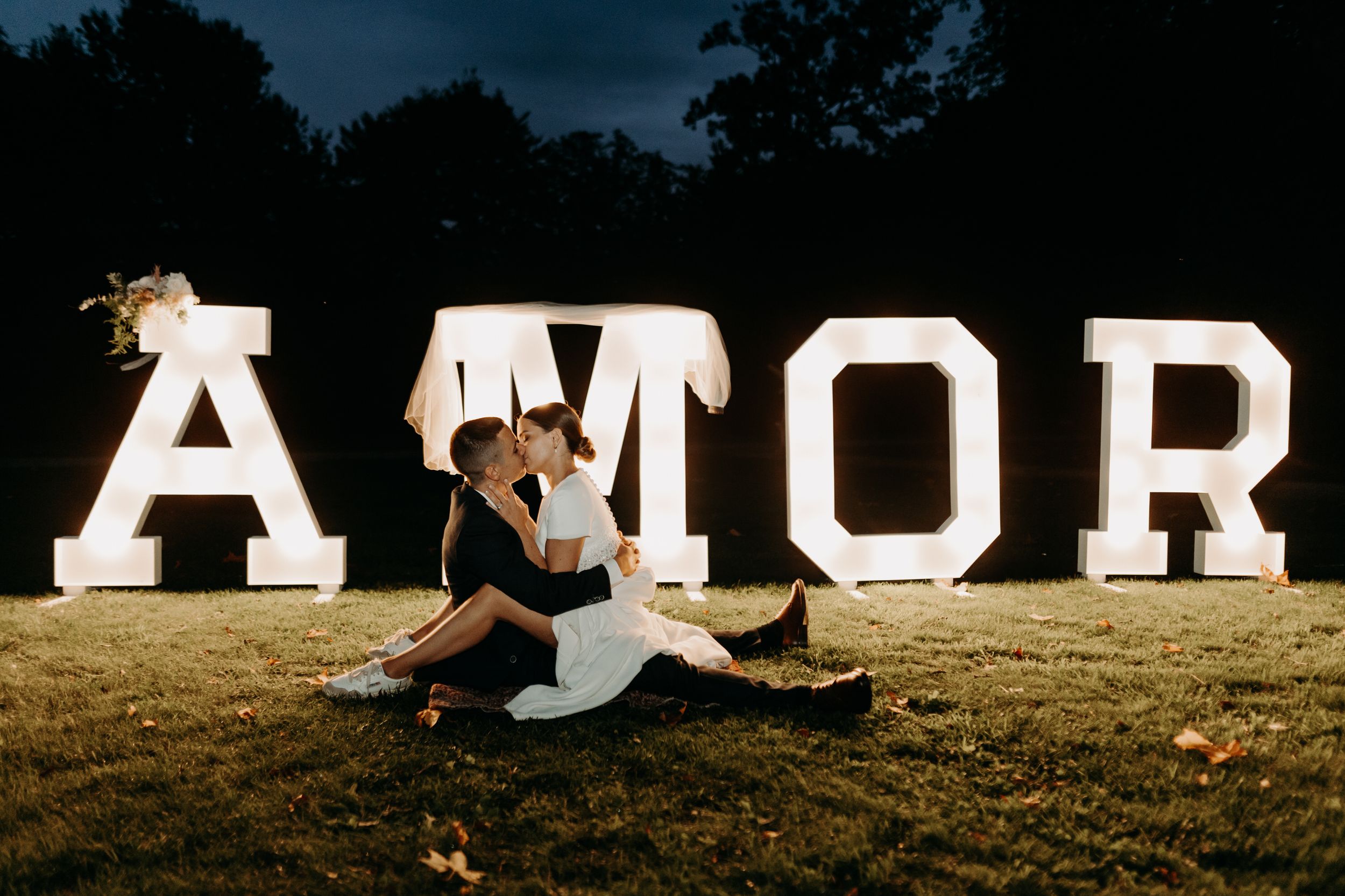 Couple seated in front of large illuminated AMOR sign at night.