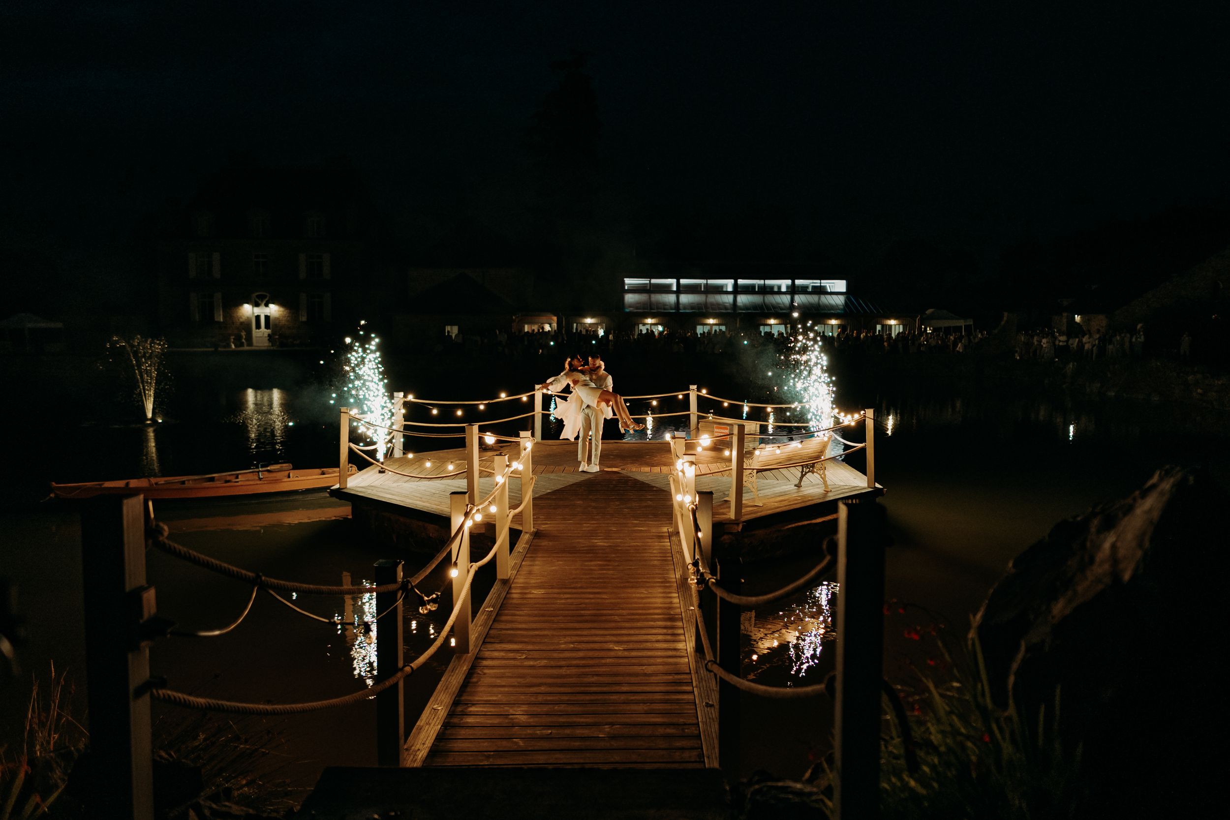 Illuminated wooden dock extends over dark water at night with lights reflecting off the calm surface.