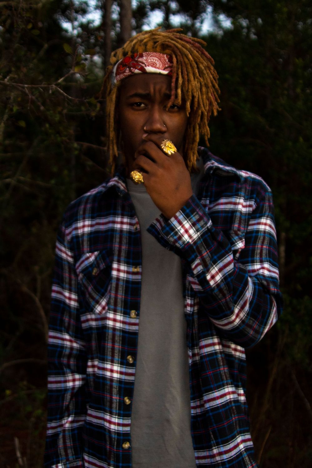 A person standing looking at the camera wearing grey and flannel shirt with their hand to their face showing their ring.