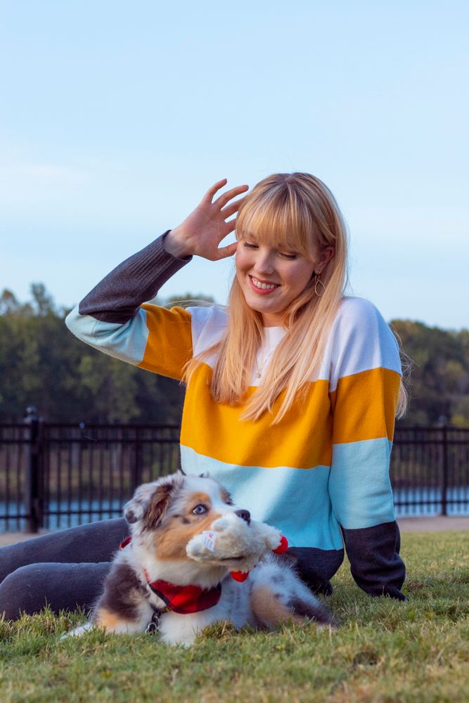 A person on the ground fixing their hair with their dog in front of them holding a toy.