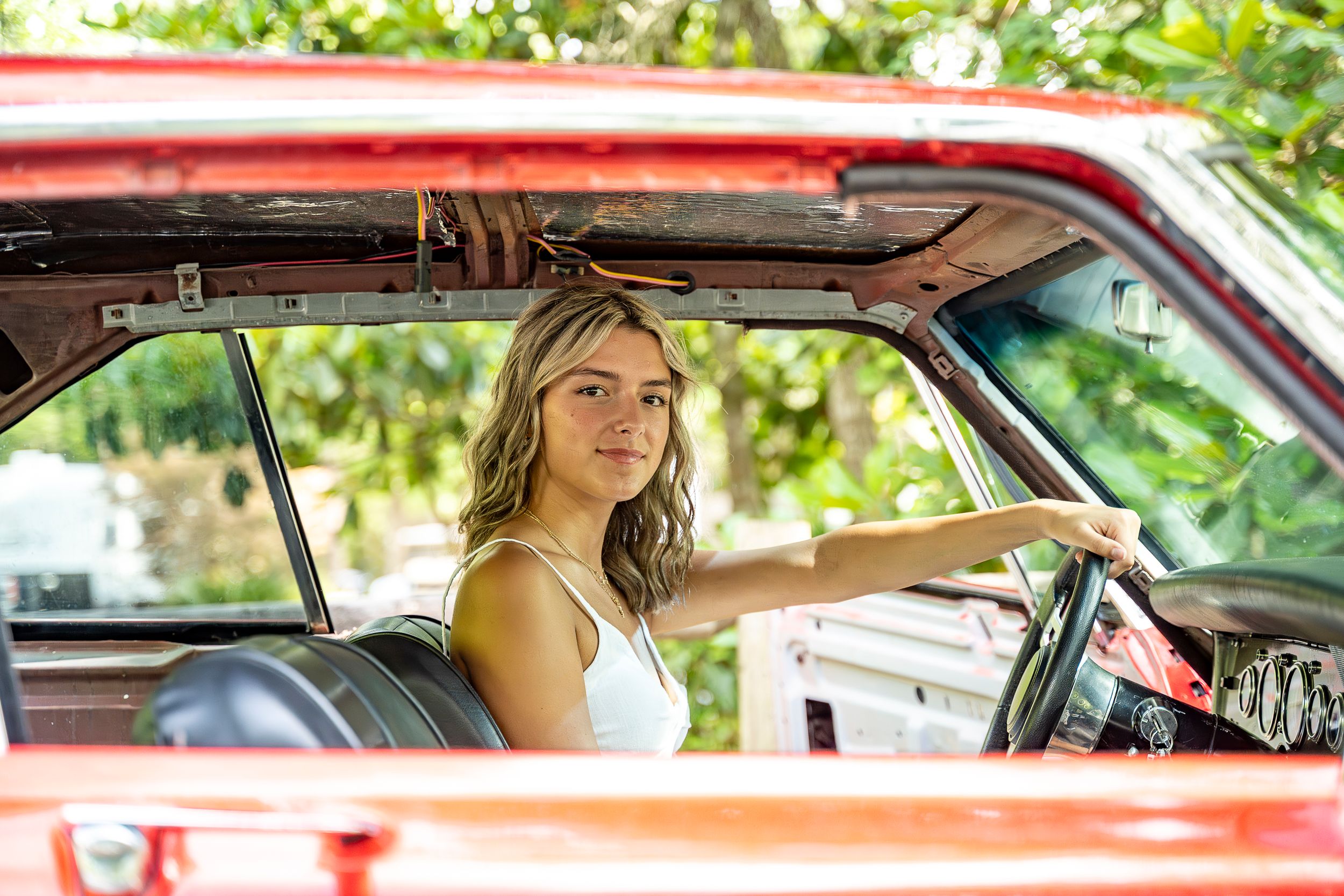 View through passenger window of vintage red car interior with sunlit greenery in background.