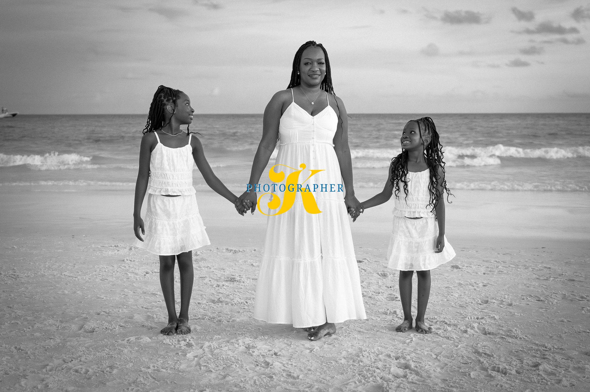 Three people in white dresses hold hands while walking along a sandy beach at sunset.