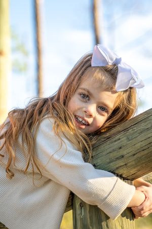 A young child wearing a white bow and sweater leans against wooden post outdoors.
