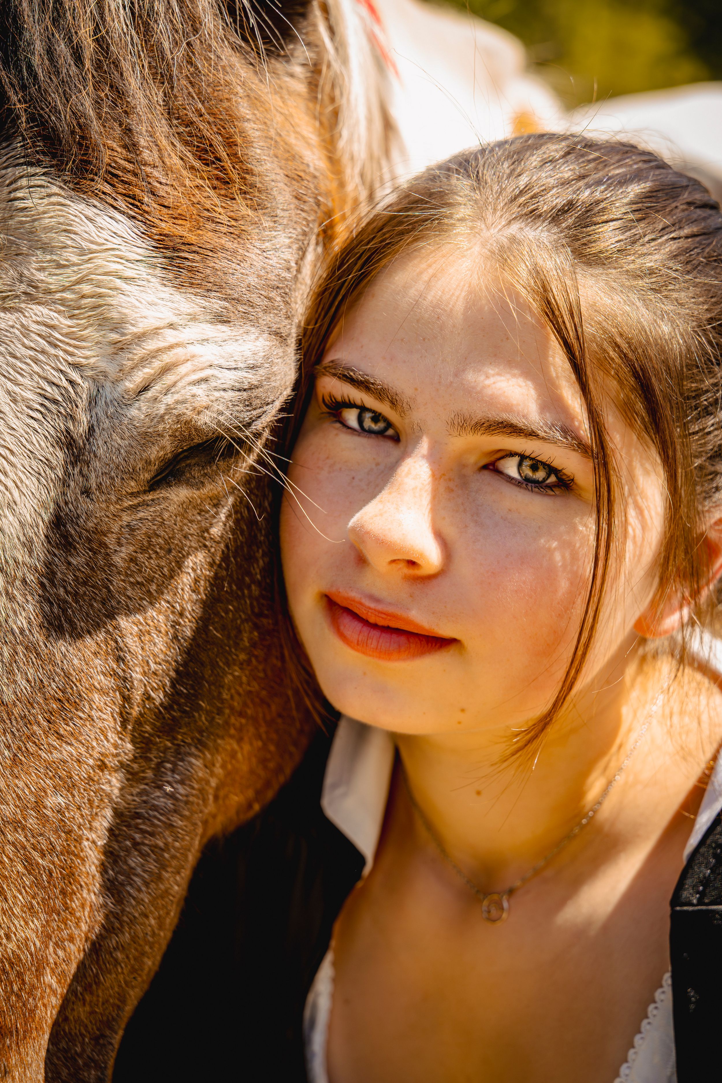 Young woman with green eyes resting her head against a brown horse, sharing a tender moment outdoors in sunlight.