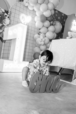 Black and white photo of one year birthday celebration with large number 1 light and balloon arch decorations.