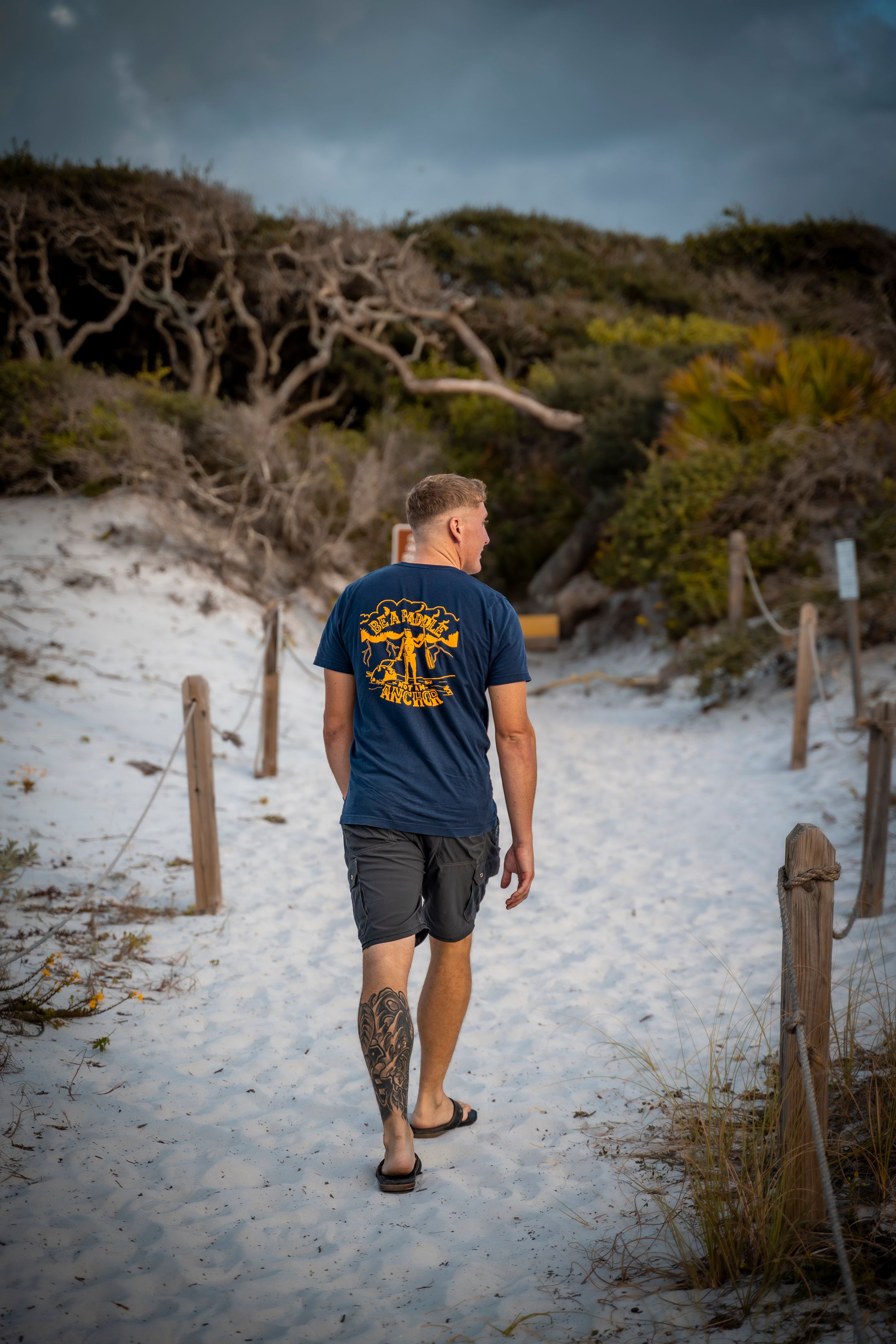 Man in navy graphic tee and shorts walks sandy beach path with dunes and trees under cloudy sky.