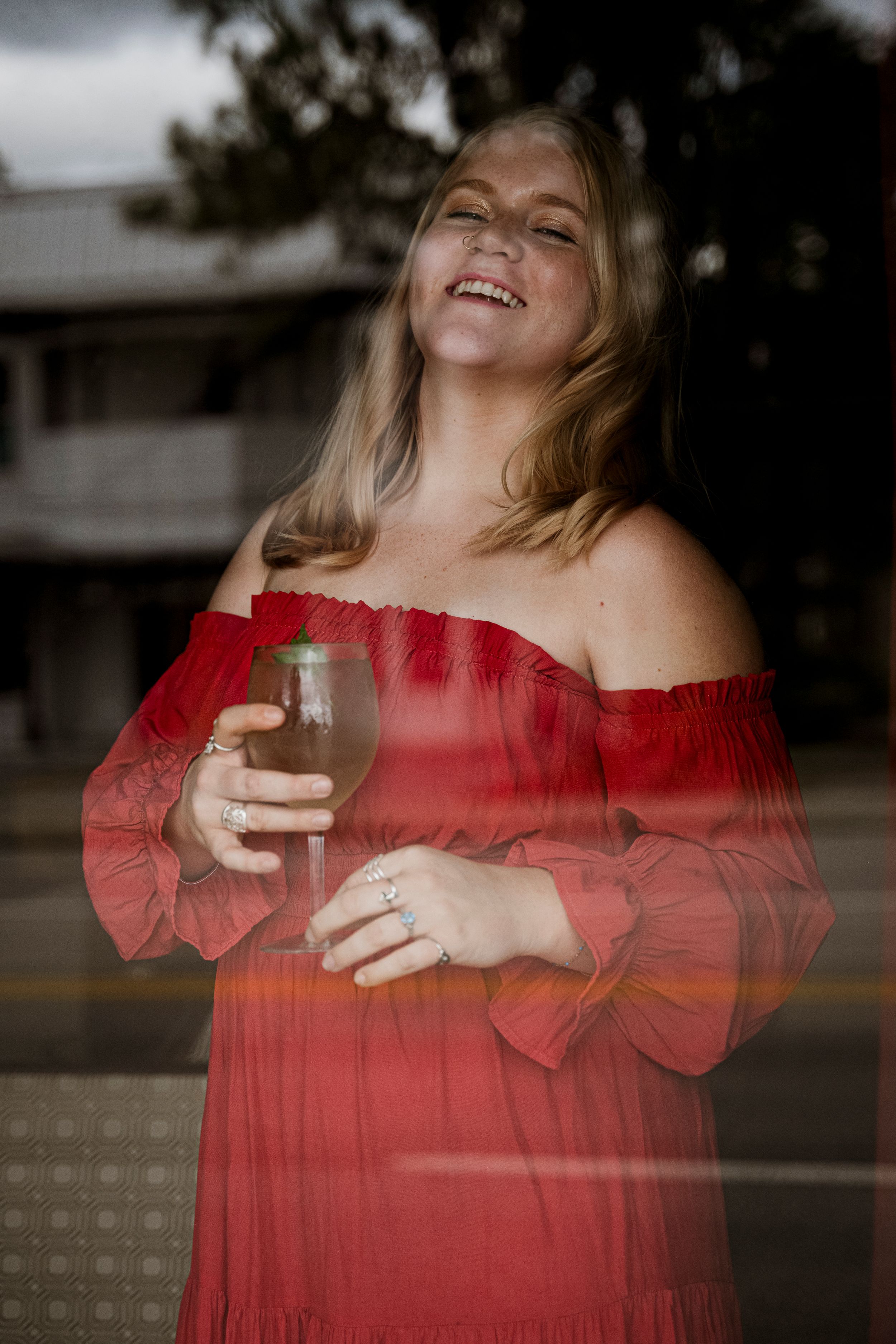 Smiling blonde woman in red off-shoulder dress holding a wine glass, photographed through a window reflection.