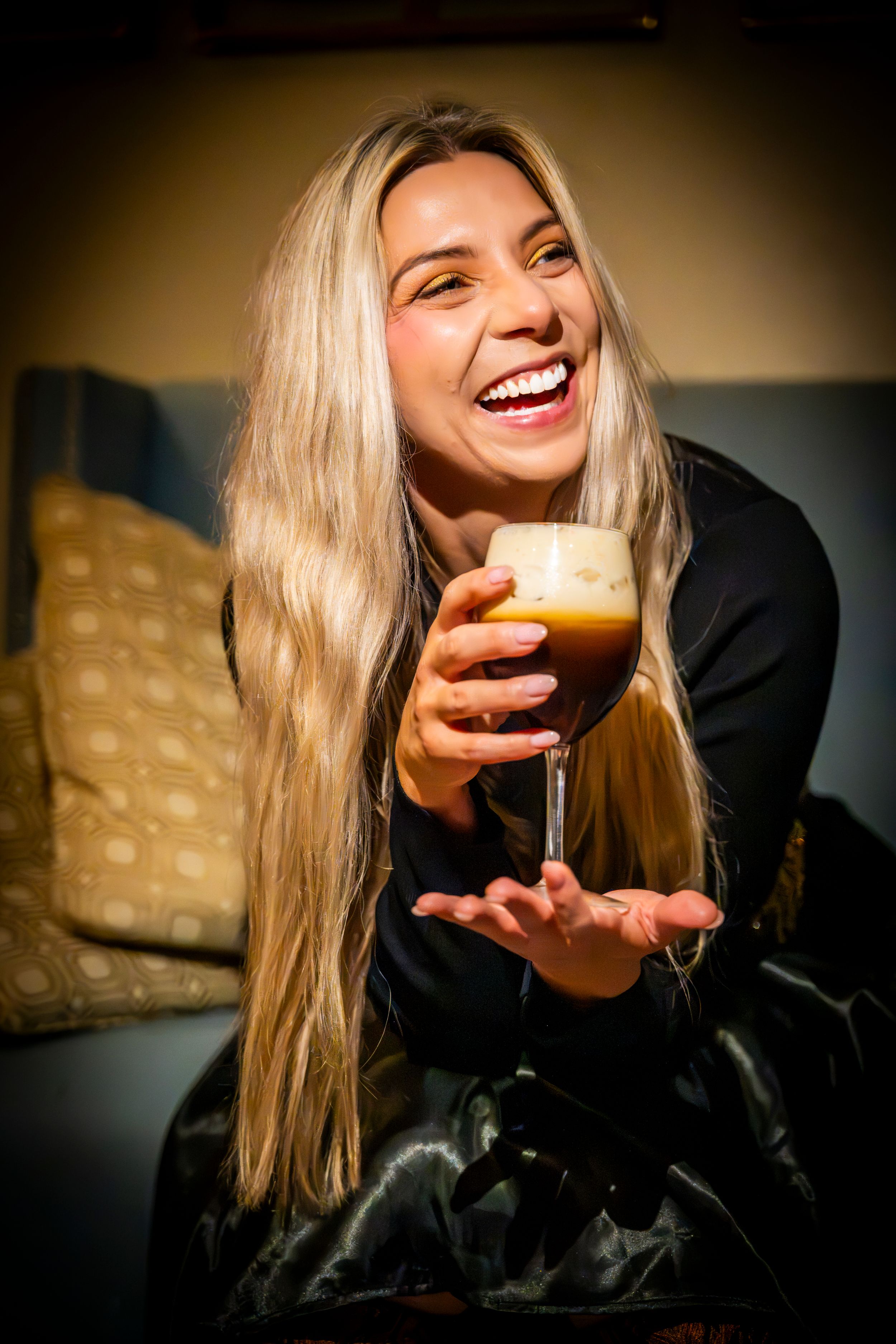 Laughing blonde woman holding a layered coffee drink in a stemmed glass while sitting on a couch.