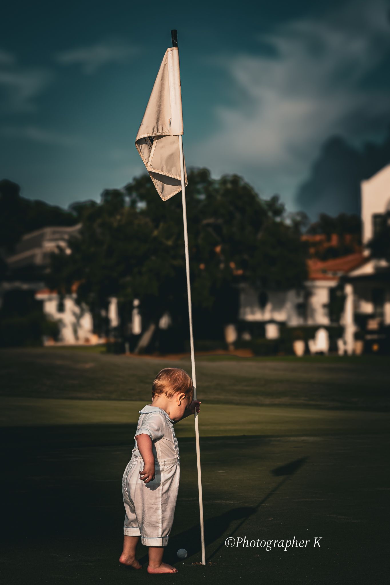 Small child in white outfit stands next to tall golf flag on green during moody evening at golf course.