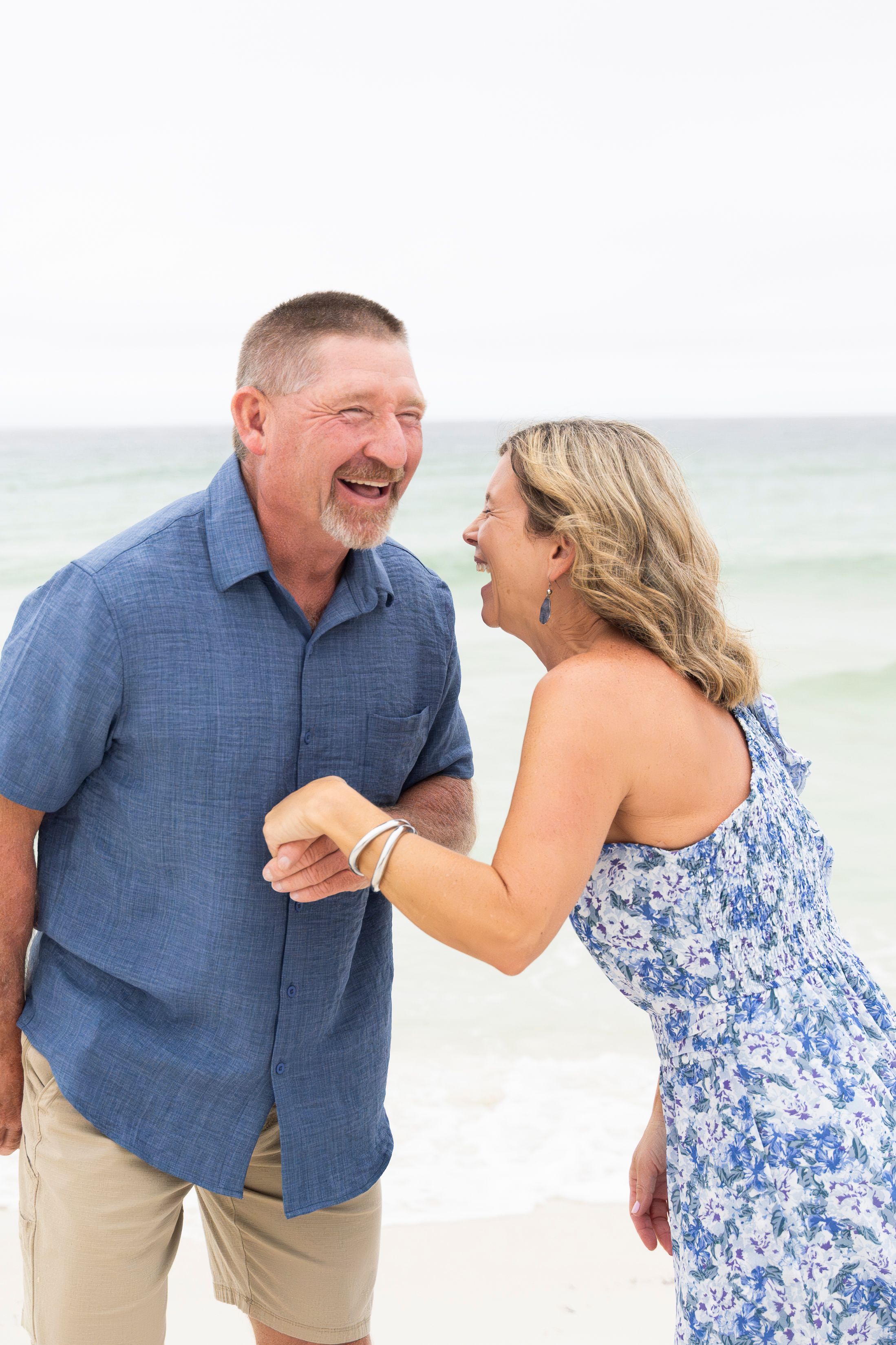 Couple in blue and khaki outfits laughing together during a beach photoshoot.