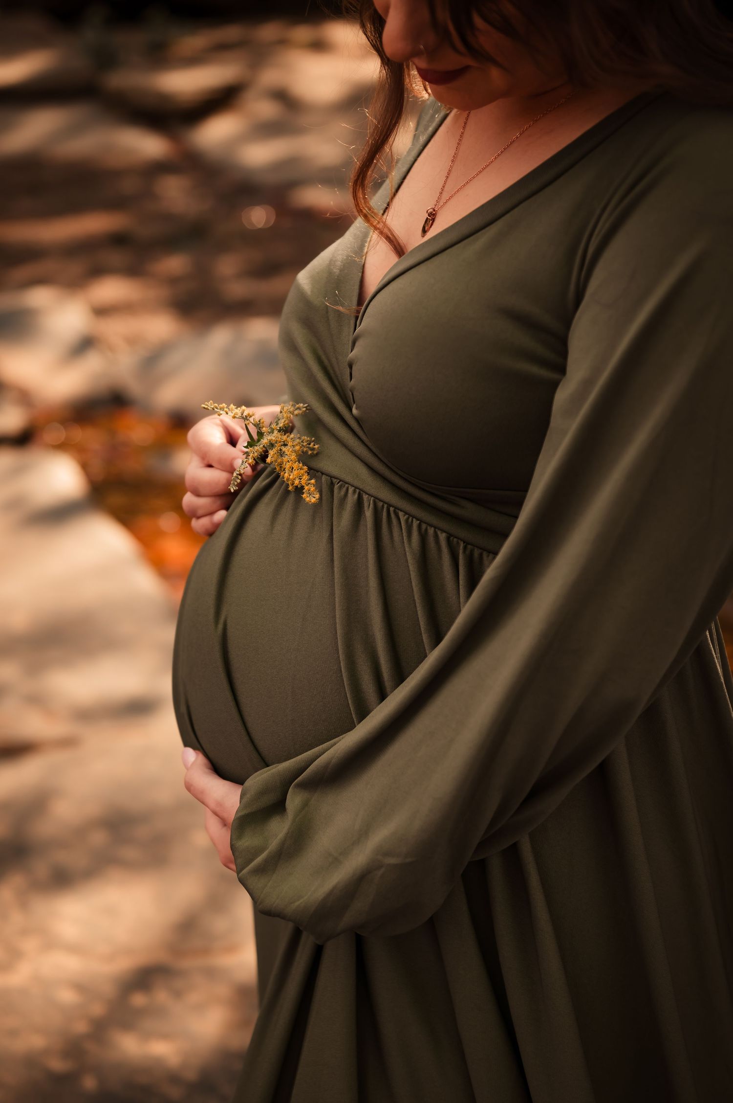 Close up of pregnant woman wearing olive green dress holding dried flowers in autumn light.