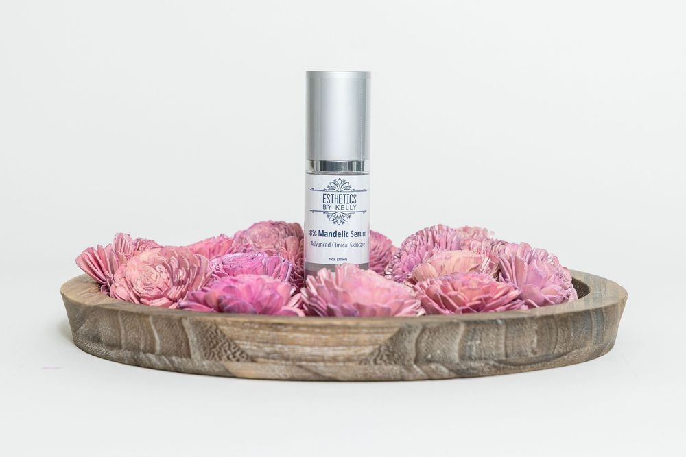 Pink peonies arranged around a silver cosmetic bottle on a rustic wooden tray against white background.