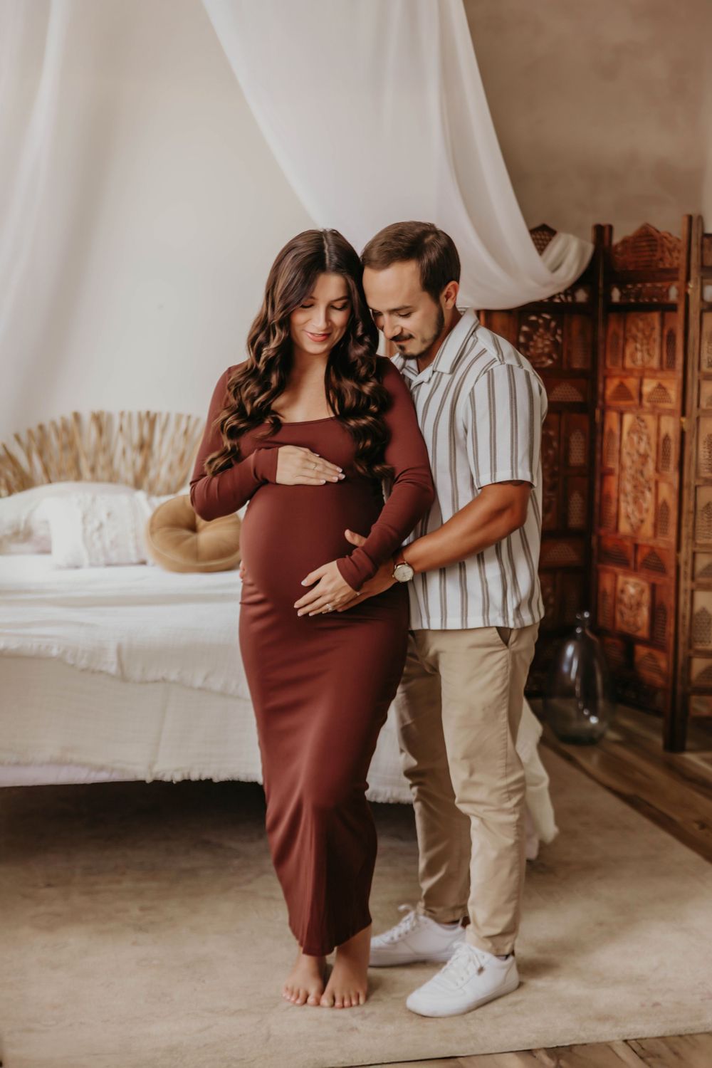Expectant couple shares intimate moment in bedroom with white drapes and wooden decor in warm tones.