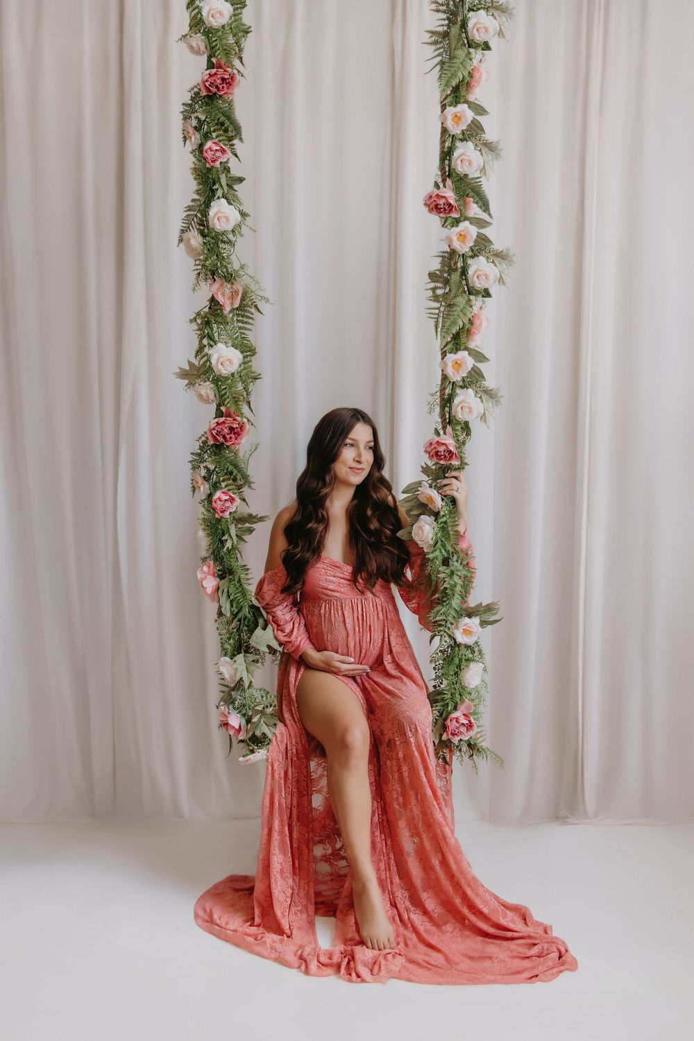 Elegant figure in flowing coral dress poses on floral swing against white backdrop with rose and greenery garlands.