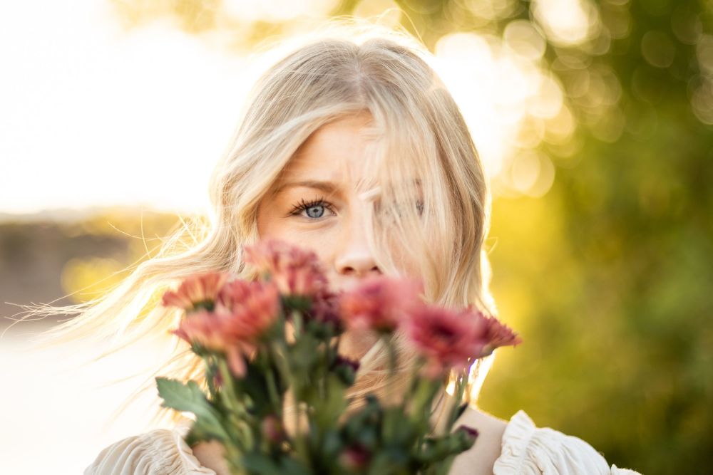 Blonde woman holding pink chrysanthemum flowers close to her face in a sunlit outdoor setting. in St. George utah