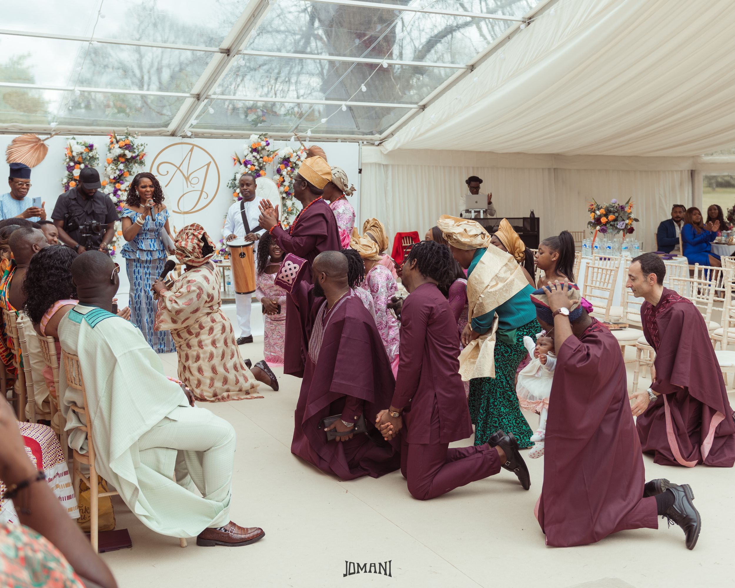 Wedding guests in burgundy traditional attire kneel during African cultural ceremony under glass marquee.