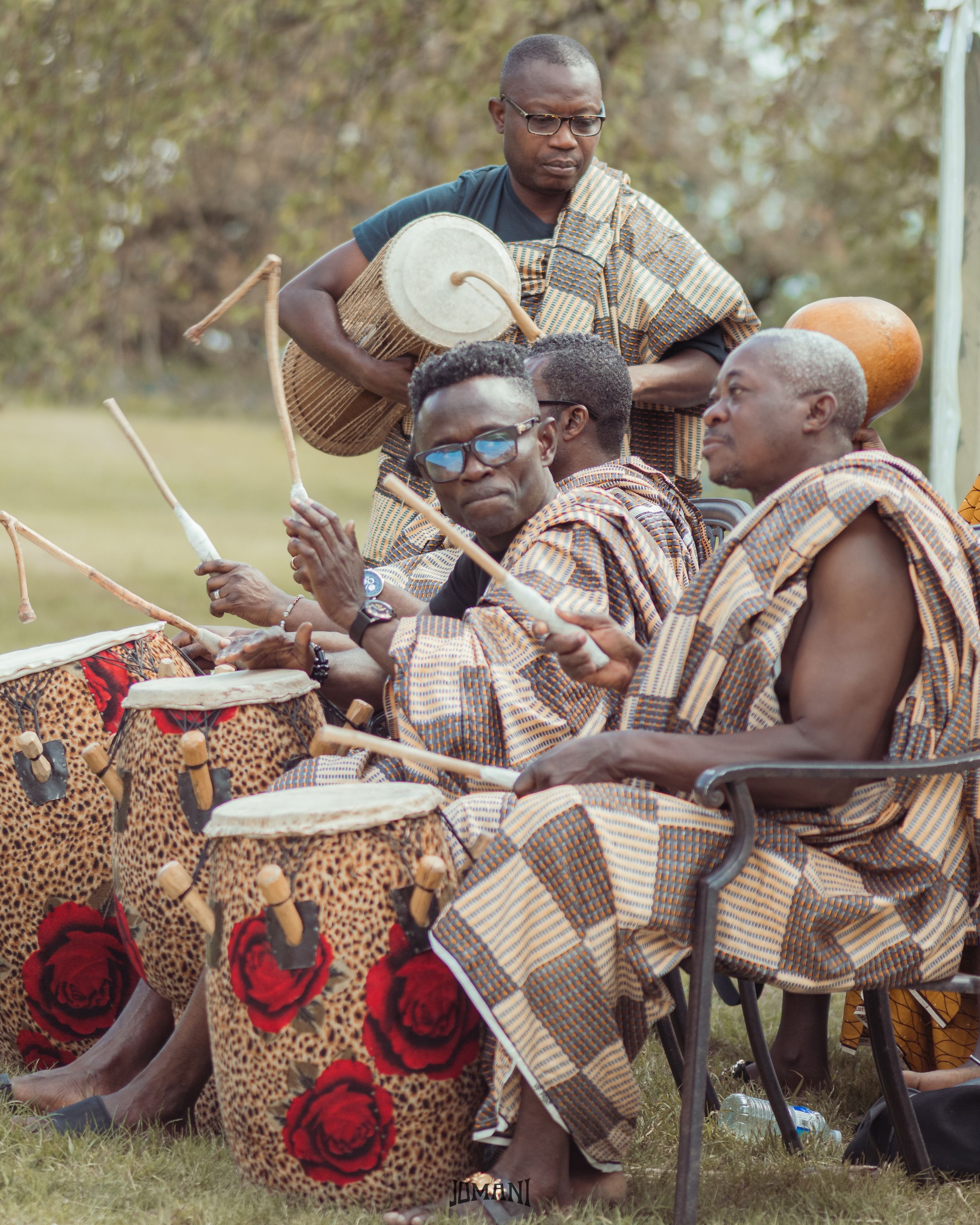 Traditional African drummers in kente cloth perform outdoors at cultural ceremony with decorated drums.