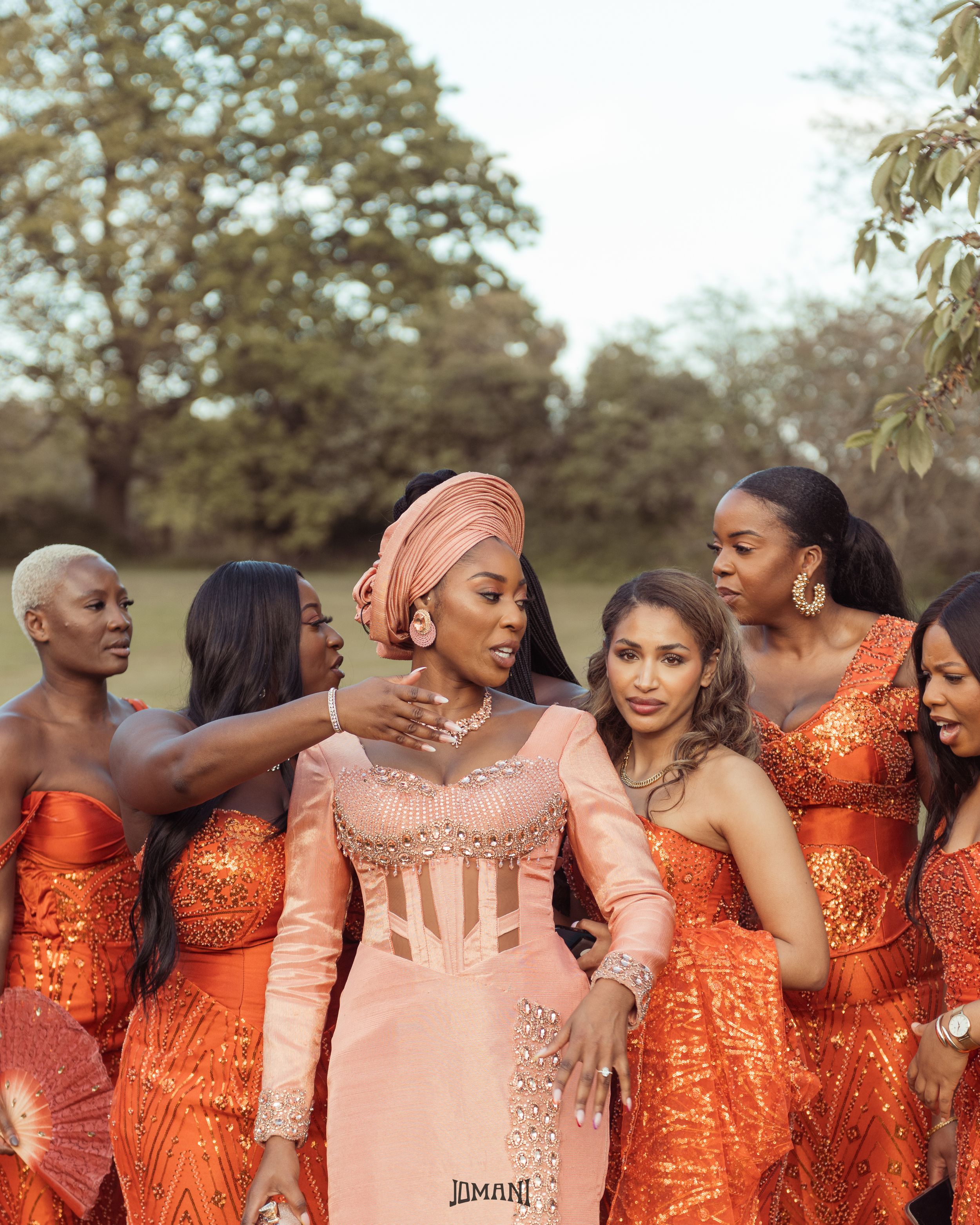 Bride in peach Nigerian lace gown with orange-clad bridesmaids outdoors.
