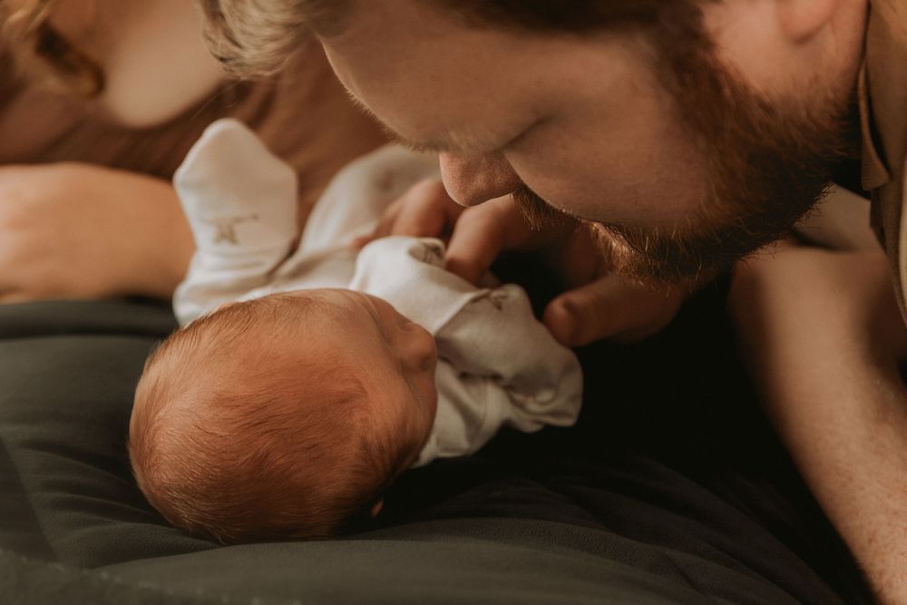 A tender moment between a parent and newborn baby lying on dark bedding in warm lighting.