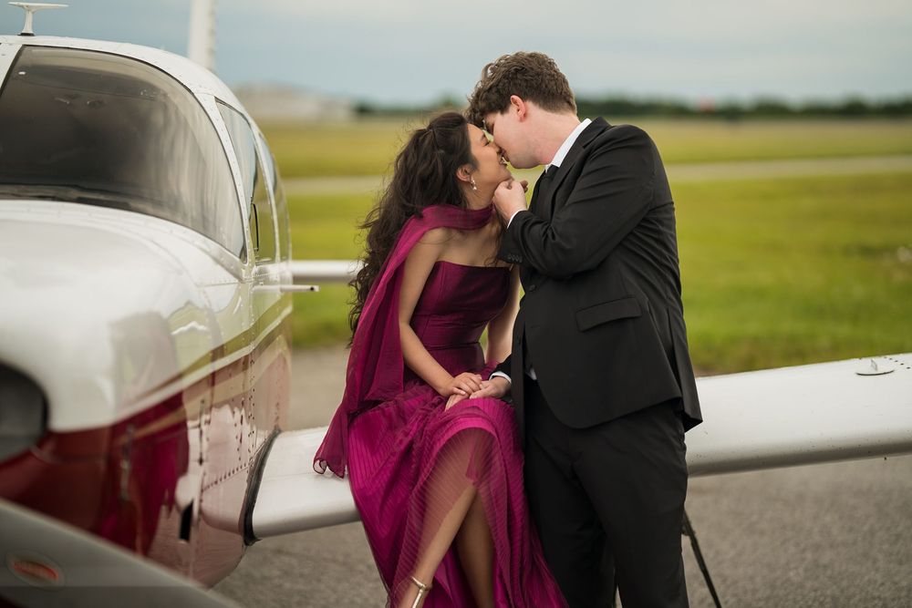 Couple in formal attire sharing a romantic kiss beside a small private plane on an airfield.