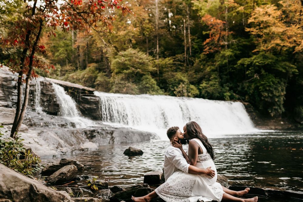 Couple sharing a romantic kiss by a serene waterfall surrounded by colorful autumn foliage in a forest setting.