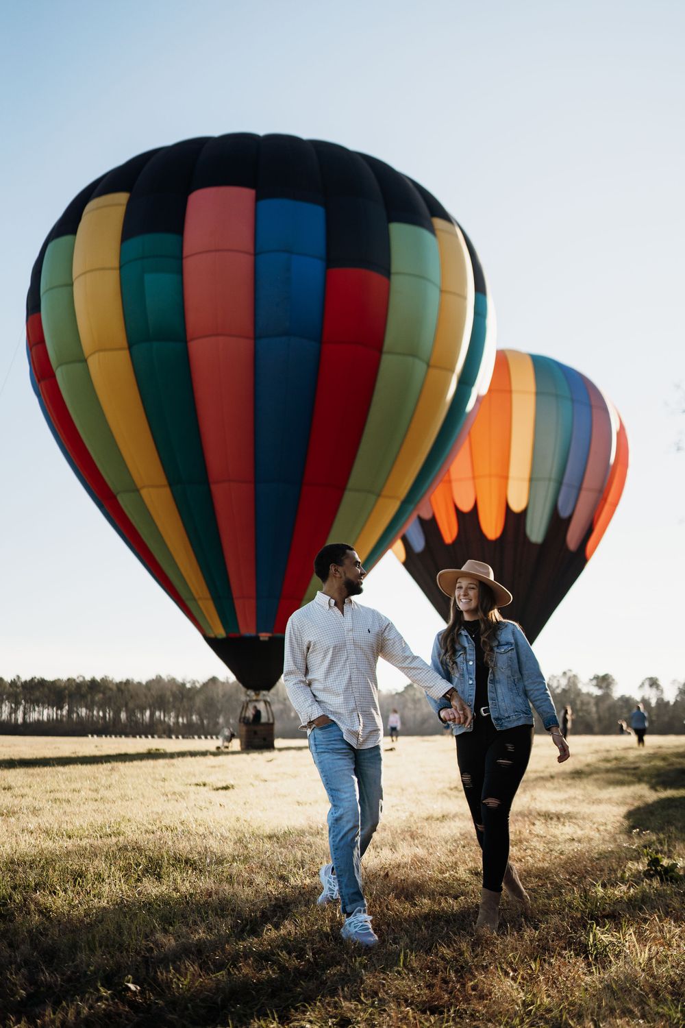 Couple holding hands walking in a field with colorful hot air balloons in the background on a sunny day.