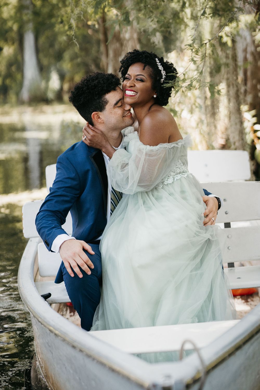 Interracial couple in wedding attire sharing a romantic moment in a white boat surrounded by lush greenery.