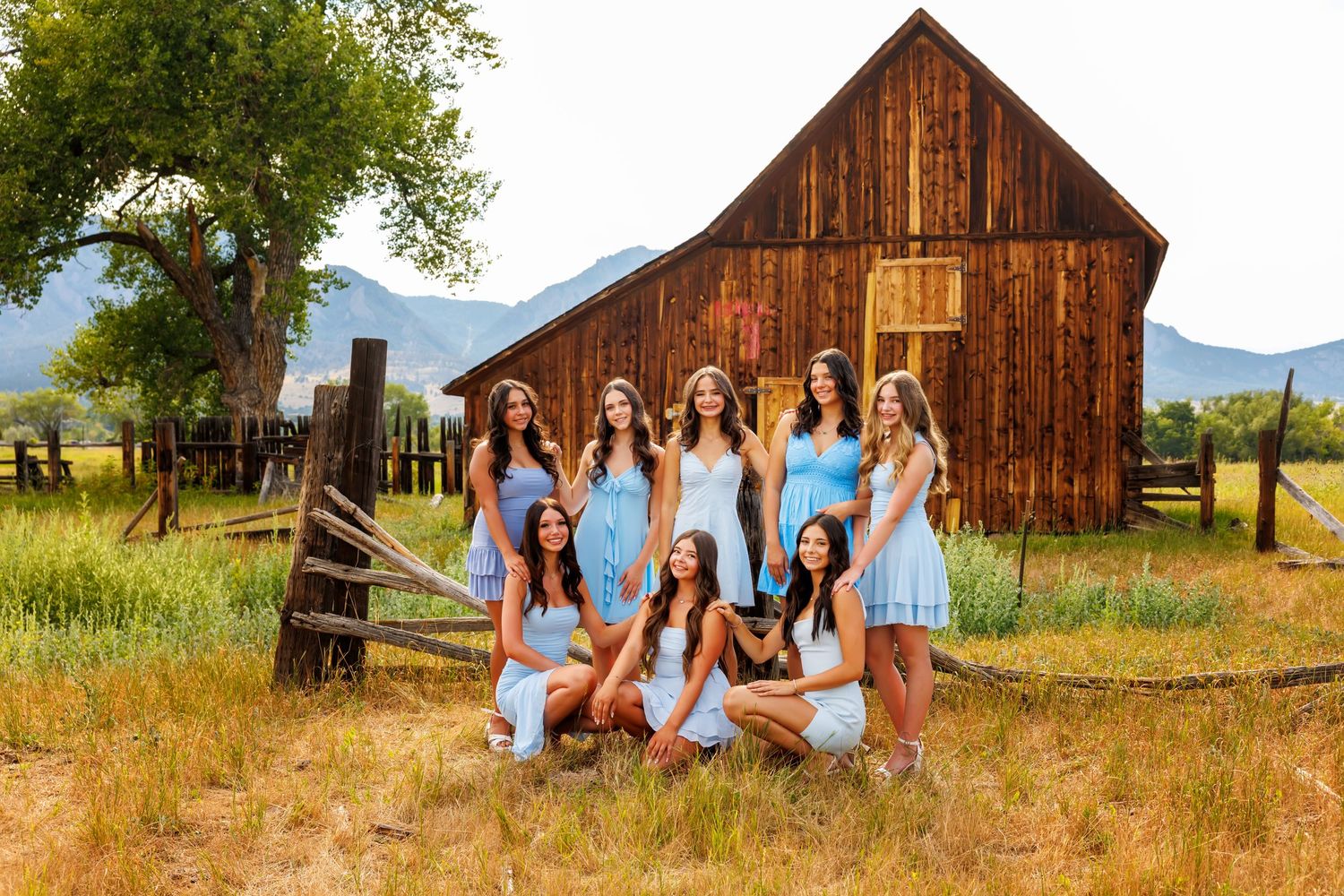 Group of young women in blue and white dresses posing near a rustic wooden barn in a scenic mountain setting.