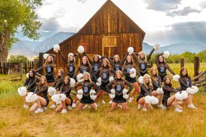 Cheerleading squad poses with pom-poms in matching uniforms before a rustic barn with mountain backdrop.