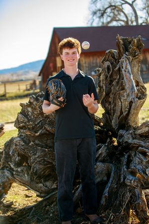 A young person stands by a large twisted tree stump holding sports equipment on a sunny day near a barn.