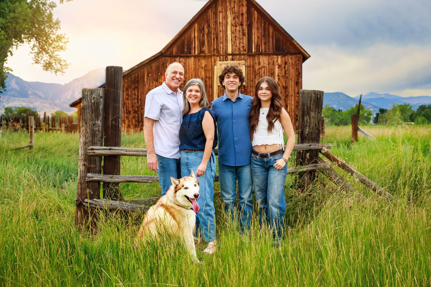 A family poses for outdoor photos in a rustic field setting with wooden barns and mountain scenery in the background.