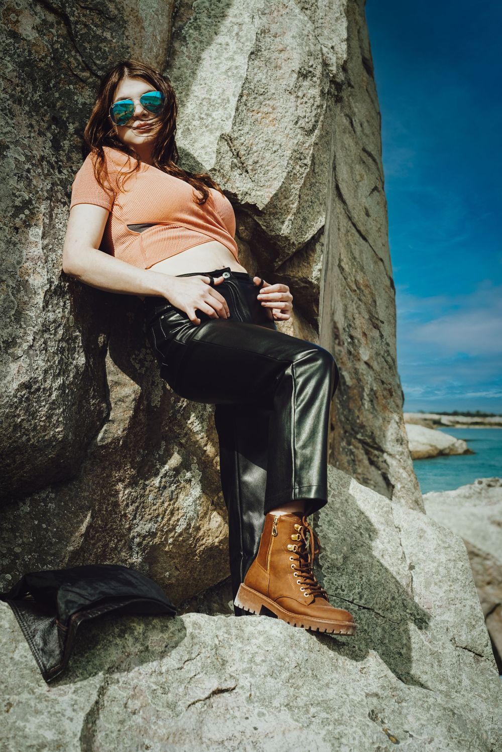 Person in orange top and leather pants sits on coastal rock formation wearing sunglasses.