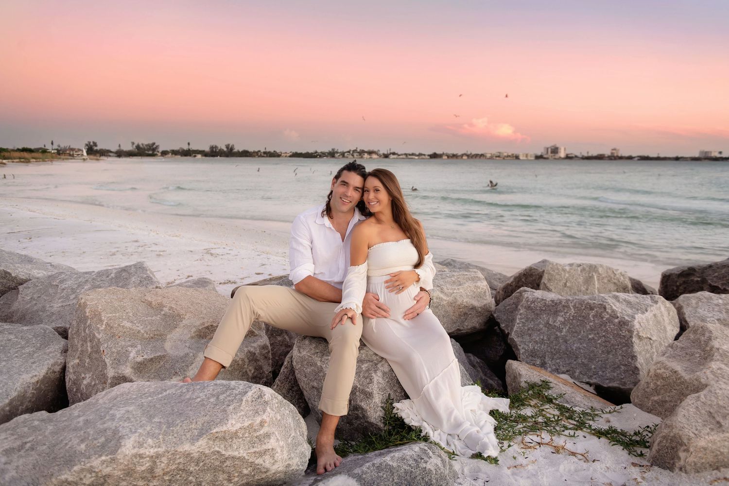Couple in white outfits sitting on coastal rocks at sunset.