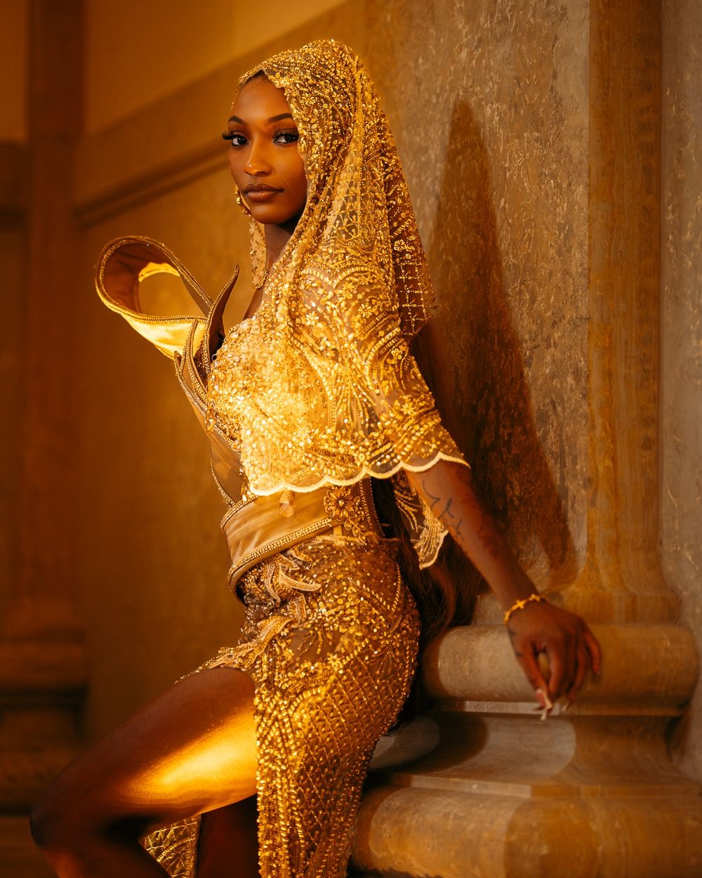Person wearing traditional golden lace veil poses in dramatic lighting against ornate architectural backdrop.