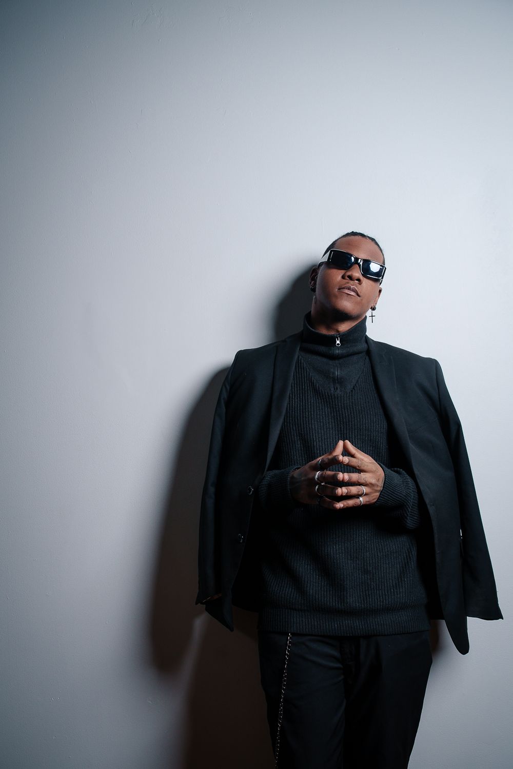A figure in a black suit and sunglasses poses expressively against a white wall in a series of dramatic shots.