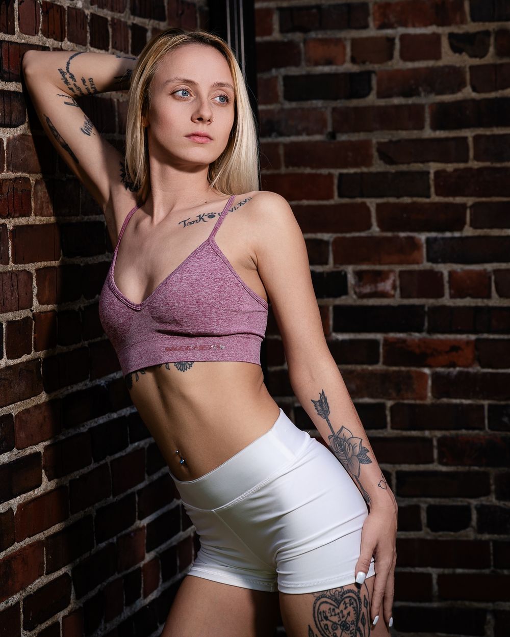 Athletic woman in purple sports bra and white shorts poses against brick wall showing tattoos in moody lighting.