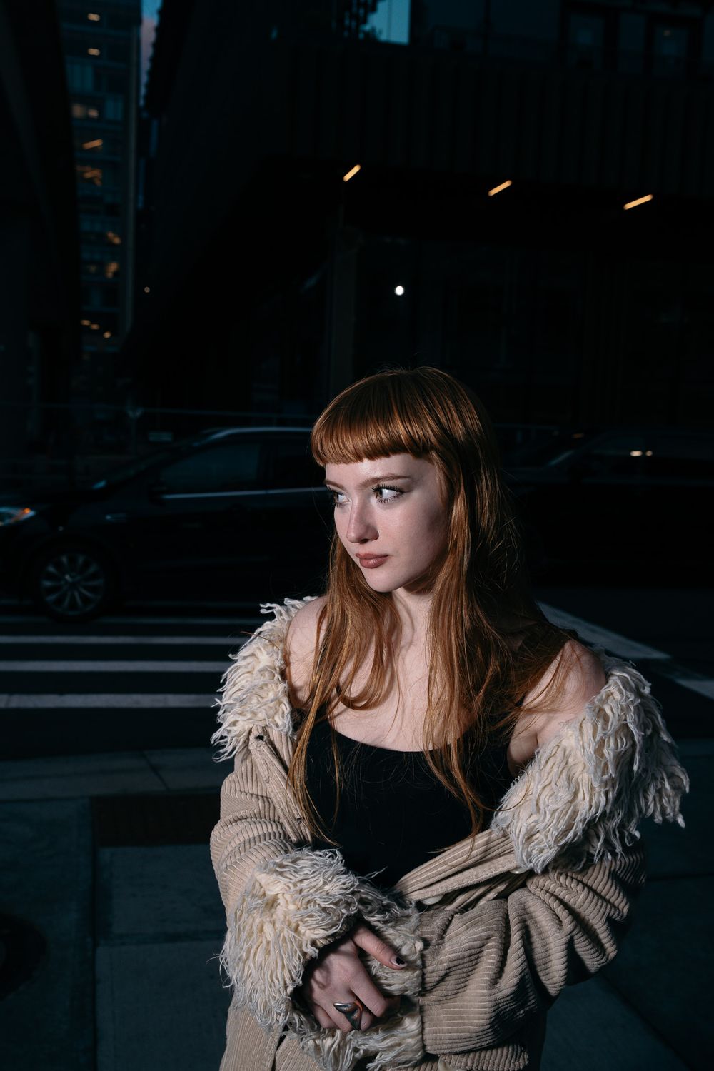 Moody nighttime portrait with model wearing fringed beige sweater in city setting.