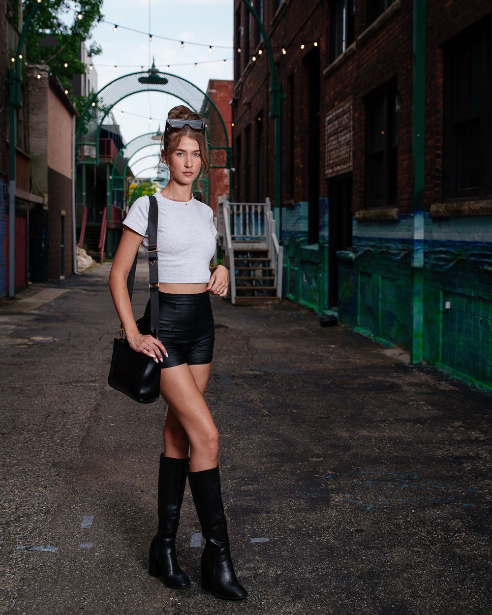 Street fashion photo showing a casual outfit with white crop top, black shorts and knee-high boots in an urban alley.