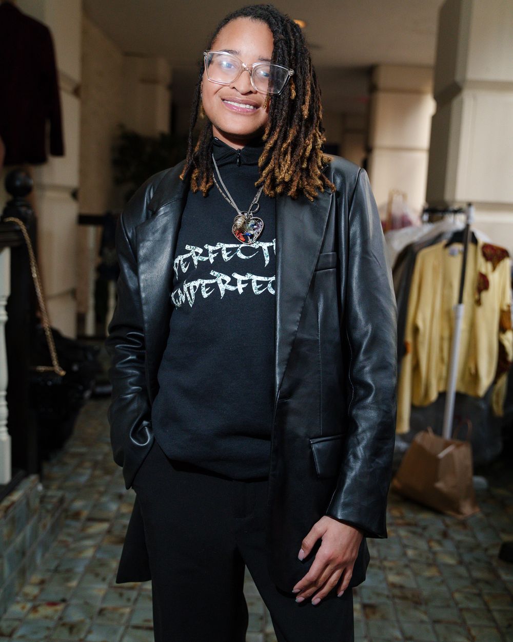 Person wearing leather jacket and graphic sweatshirt with dreadlocks standing indoors.