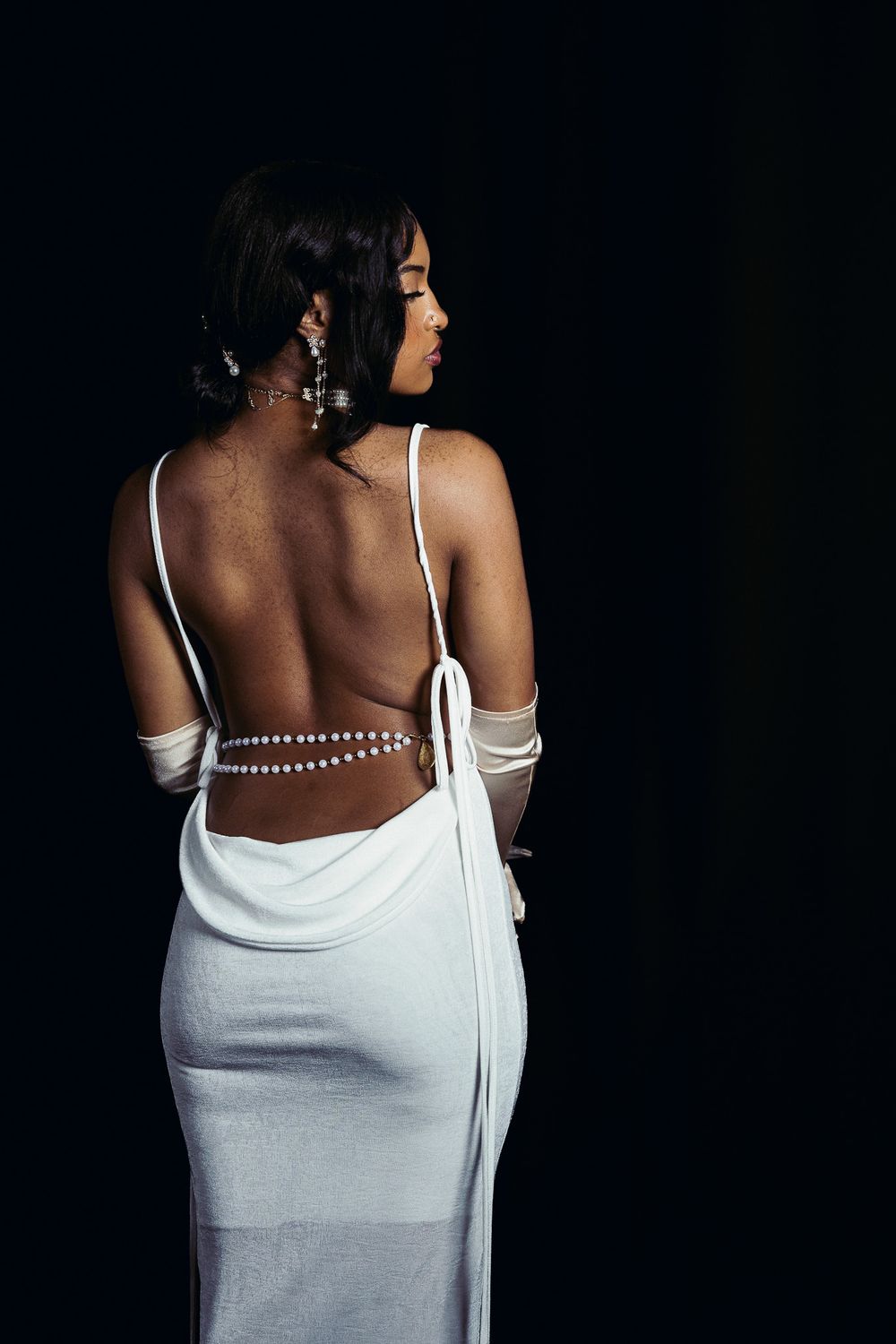 Back view of white evening gown with pearl straps and open back design against black background.