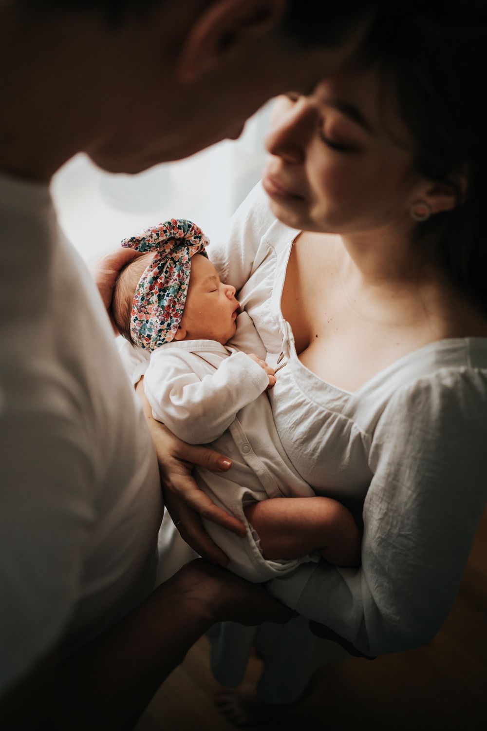 tata i mama stoją przy oknie, stykają się czołami, między nimi noworodek - sesja noworodkowa Kraków