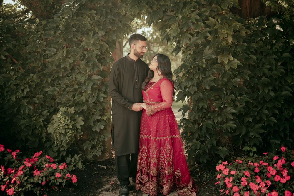 Couple in red and black traditional outfits posing in garden with blooming flowers.